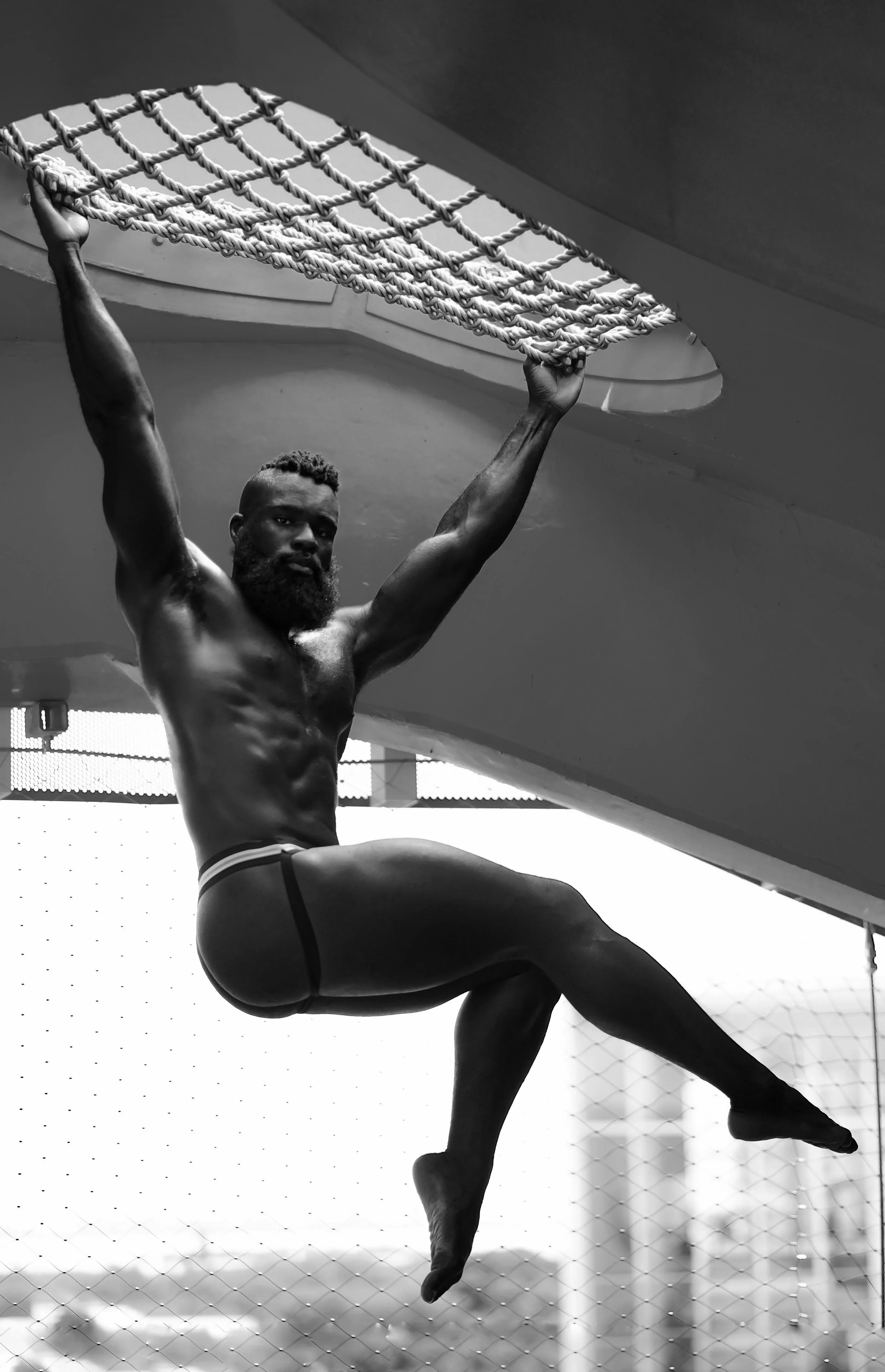 Athletic man in black jockstrap underwear hanging from rope net indoors, strong physique
