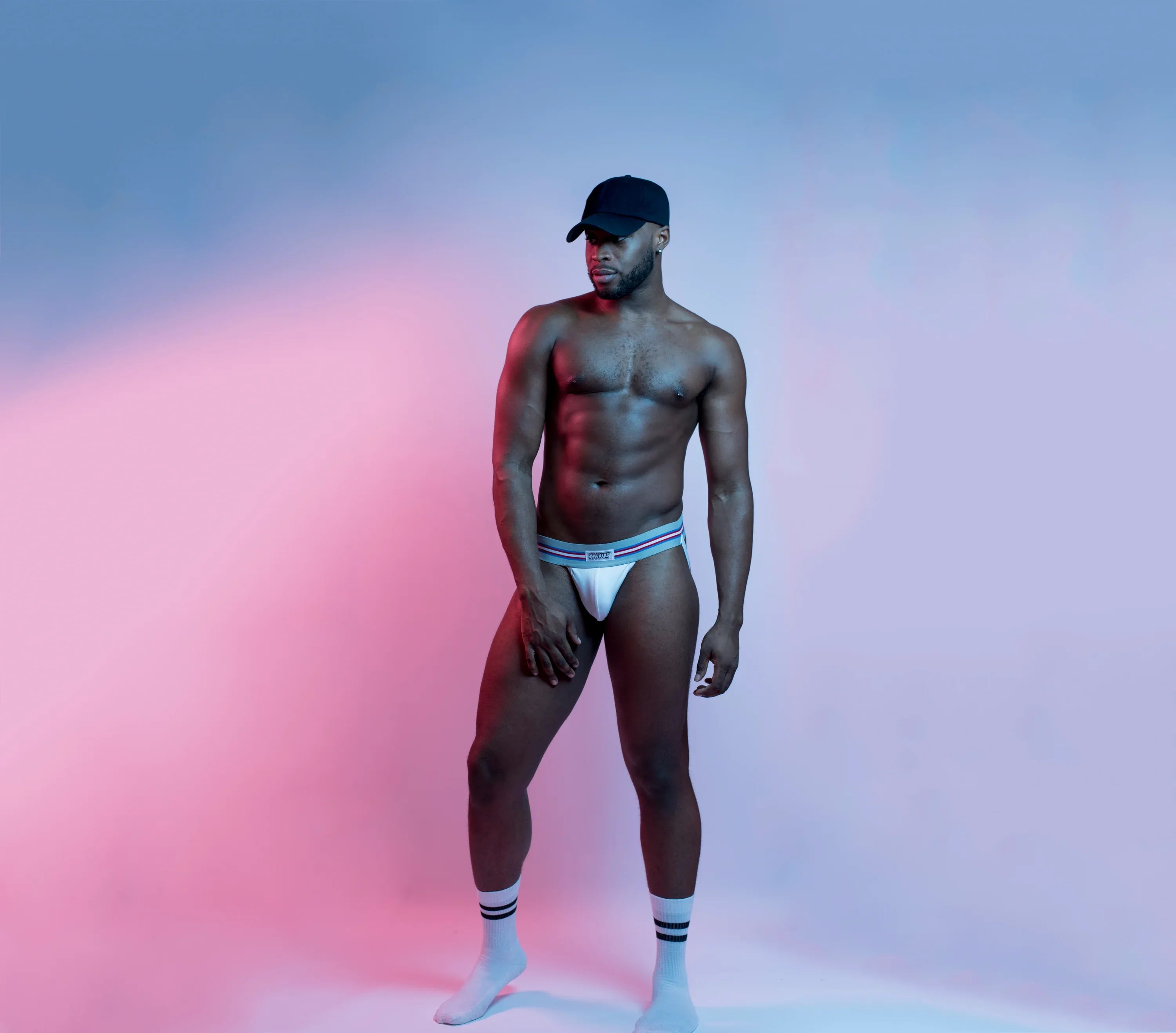 Athletic man modeling white men's jockstrap underwear, socks, and cap in studio lighting